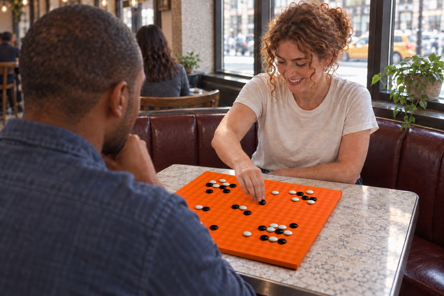 Illustrative scene of two people playing Go on an orange Sine board at a café table. The full photograph is shown.